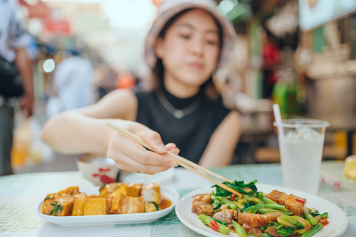 Singapore vacation asian woman smiles teasingly as she sits with her friend at a table in a sidewalk cafe in Singapore's Chinatown. eating rice with asian street menu fried tufu and grill pork with vegetable