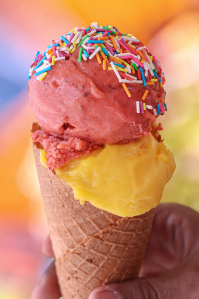 istock Close-up image of unrecognisable person holding waffle cone, two scoops of mango and strawberry gelato ice cream, multicoloured sugar sprinkles, vibrant coloured background, focus on foreground 2150499072