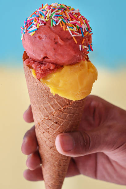 istock Close-up image of unrecognisable person holding waffle cone, two scoops of mango and strawberry gelato ice cream, multicoloured sugar sprinkles, blue-yellow split background, focus on foreground 2150499092