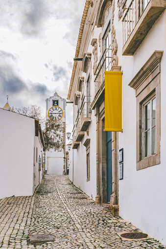 A cobblestone alley in Tavira, Portugal, leading to a tall clock tower, with ornate facades and yellow banners along the buildings.