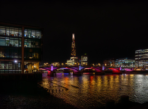 Blackfriars Bridge illuminated at night with purple lights, reflecting its lights on the River Thames and its bank, with the Shard skyscraper behind and illuminated office buildings. London.