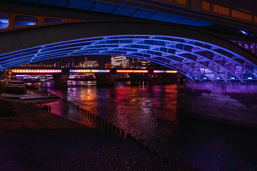 Steel structure of the lower part of a London Bridge illuminated in blue and purple colors reflecting its light on the River Thames and its bank in London.