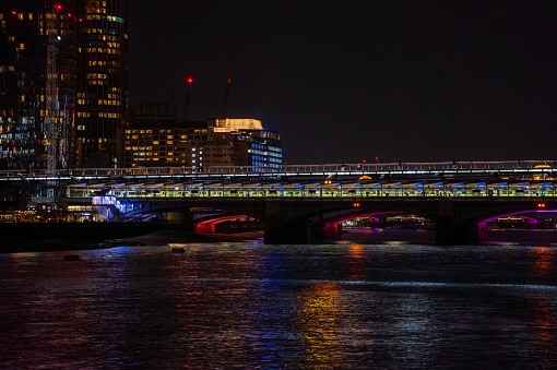 Night train traveling over Blackfriars Bridge in London at night, with the purple lights reflecting in the water of the River Thames and the Millennium Bridge in the background.