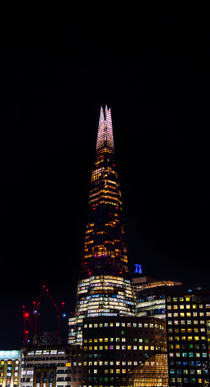 Night view of the London skyscraper The Shard with the sky completely dark and buildings below with the windows illuminated in many colors.