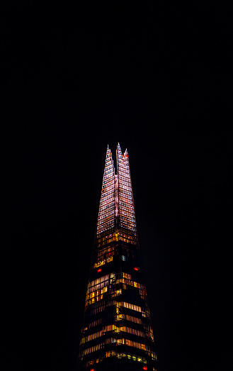 Above and top of the London skyscraper The Shard with the sky completely dark at night with its illuminated metal penthouse structure and illuminated windows in many colors.