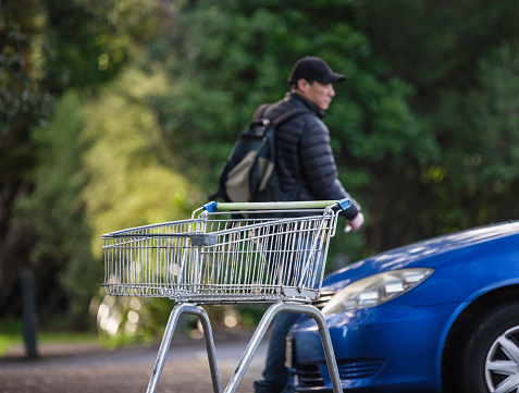 An empty supermarket shopping trolley dumped at the carpark. Man walking towards his car.