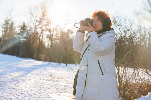 Caucasian ginger haired woman photographer in earmuffs, is capturing sunny winter park. Back lit and sunlight effect