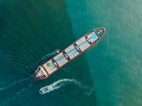 The tug accompanies the ship when entering the cargo port. 
Aerial drone view above sea port. Cargo vessel is loading containers and bulk. Professional business logistics and transportation of cargo ship.