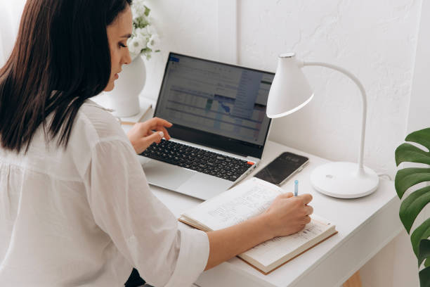 istock Back view of young freelancer woman sitting at white desk using laptop writing notes while studying online, watching webinar, looking at pc screen learning web classes or remote working from home 2150520203