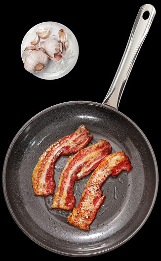 Traditional fried juicy crunchy gourmet bacon rashers in a large new modern heavy duty black frying pan, with non-slip ceramic double coated inner surface, and pair of Garlic bulbs with few detached cloves on white rimmed porcelain plate set beside, isolated on black background, viewed directly above, high resolution stock image.