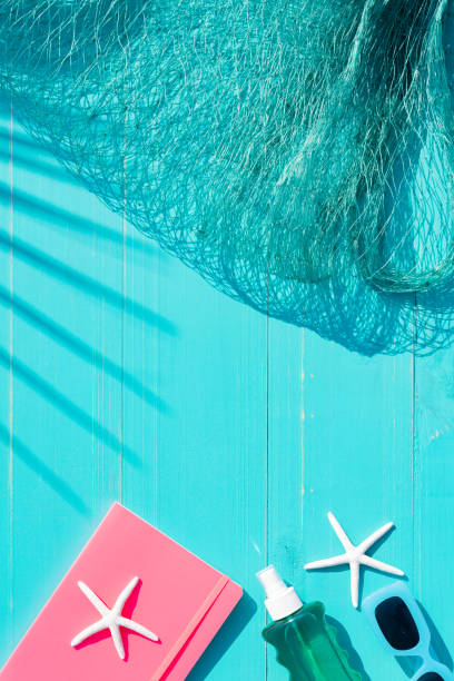 Vertical composition view from above with turquoise blue background on wooden boards with pink book, sunglasses, fishing net and palm leaf shadow vacation concept.