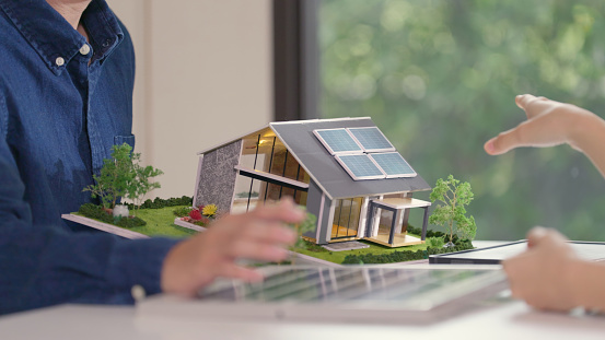 An expert gestures towards a house model with solar panels, outlining the environmental and economic advantages of solar power to a pair of potential homeowners.