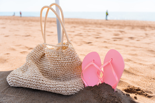 Close-up of a beach bag next to a flip-flop and an umbrella on a bright summer day and an out-of focus person in the background