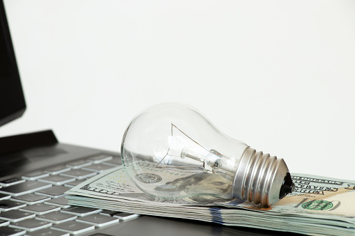 Incandescent lamp lies on dollars on laptop, high price of electricity, buy light online