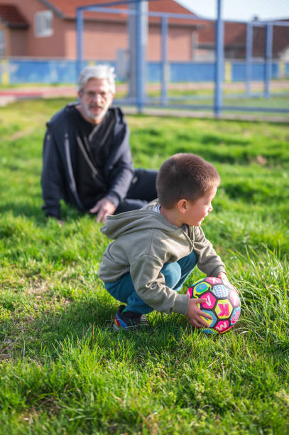 Image shows a 3 year old toddler standing by a soccer ball near a lying grandfather, symbolizing bonding, early football interest, and family playtime
