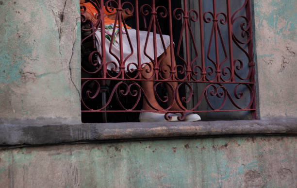 An old woman wearing a traditional dress sitting at her window in front of a window grate with only her legs visible.