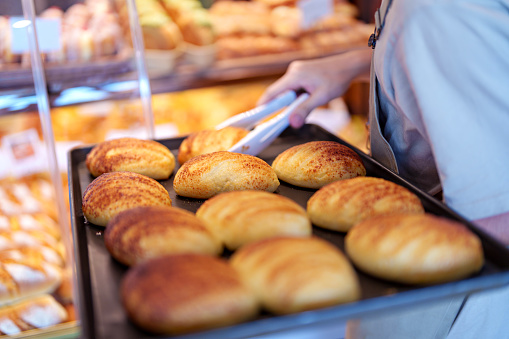 In the cropped shot, a bakery owner is seen refilling a tray of freshly baked bread buns in the display rack, ensuring that customers have access to delicious treats.