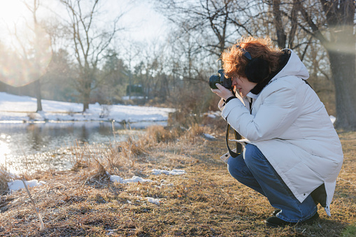 Caucasian ginger haired Caucasian woman photographer in squatting position, takes photos on the lake bank in a sunny winter park. Profile view