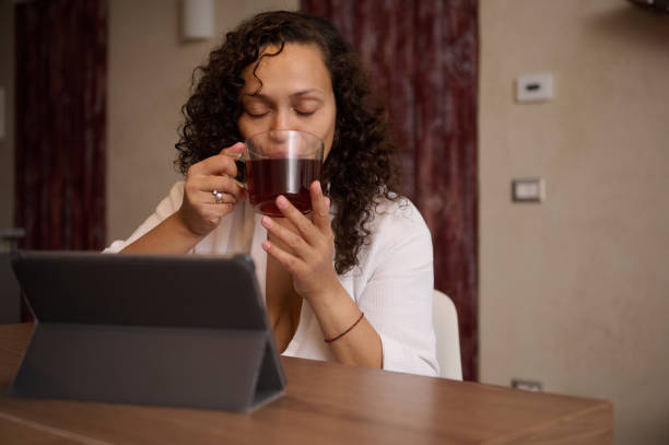 istock Beautiful multi ethnic middle aged woman in white bathrobe, drinking hot tea, sitting at digital tablet at home interior 2150543392