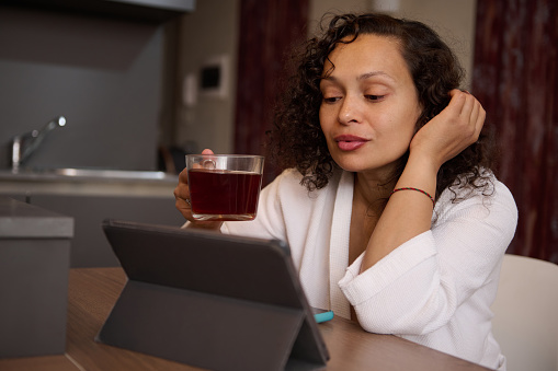 Attractive delightful young brunette woman reading news on digital tablet, browsing web sites, sitting at kitchen table over a cup of tea, dressed in white bathrobe