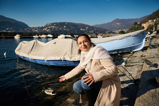 Attractive delightful young adult woman in beige coat, sitting nears boats by the Como lake, relaxing in the lakefront, smiling looking at camera while feeding by hands the birds and ducks with bread