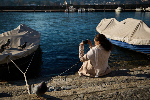 Back view of a young woman in beige coat, sitting nears boats by the Como lake and taking photos of the mesmerizing landscape on her smart mobile phone. Pretty female tourist relaxing in the lakefront