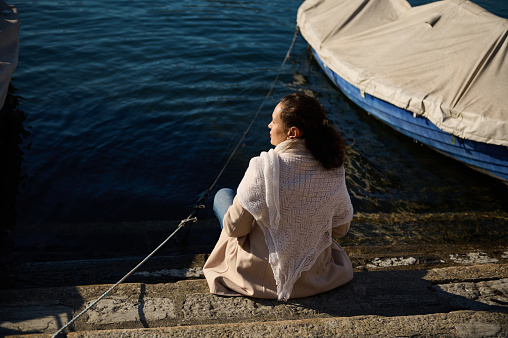 Rear view of a young woman relaxing, sitting near small boats on the edge by lake, admiring the beautiful Alpine nature from the waterfront. Como, Milan, Lombardy, Italy. Travel and tourism concept.
