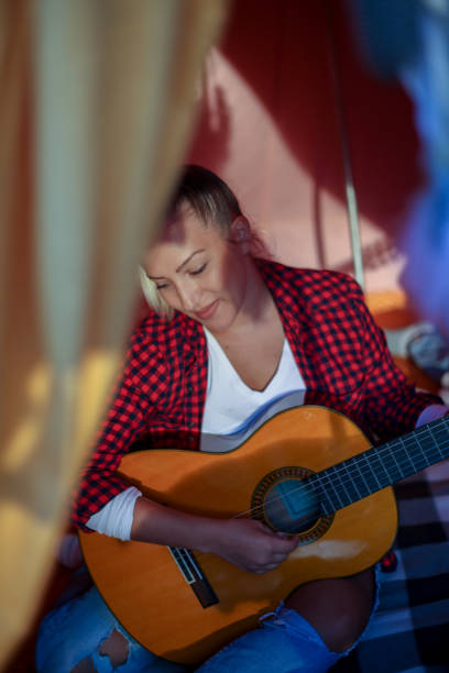 Witness the beauty of a musical interlude in the midst of nature as a young woman sits by her tent, playing the guitar, her music blending seamlessly with the peaceful ambiance of the surroundings