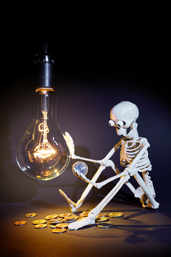 A toy human skeleton sits next to a large, dimly lit light bulb, he is surprised by the high cost of electricity.