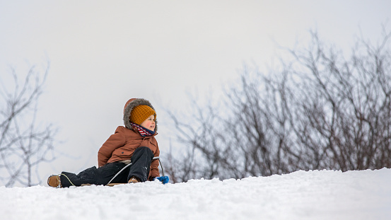 A smiling boy who has sledded down the mountain straight into the deep snow is sitting on the snowdrift and looking back for help.