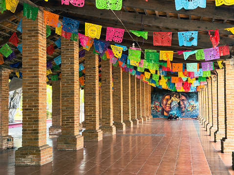 San Pancho (San Francisco), Nayarit, México - 2024-04-12: Colorful papel picado banners crisscross the Plaza del Sol in San Pancho, where a mural of embracing figures adds a vibrant cultural touch to the public space, reflecting the artistic spirit of the community.