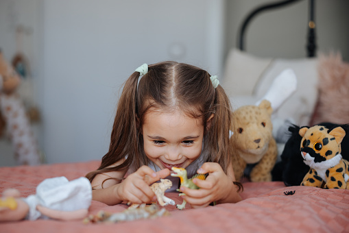 Cute Little Girl Blond Laying on a Bed at Home, Plays with Toy Dinosaurs. Happy Child Playing with Toys in Sunny Bedroom. Close-up Portrait Shot.