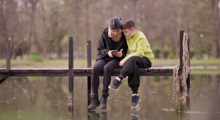 Friends enjoying a selfie on a peaceful lakeside dock. Mother and son taking a selfie while sitting on a dock overlooking a serene lake, encapsulating a moment of joy and companionship in nature.