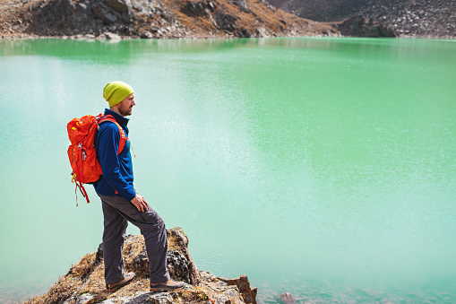 Mia adult man with backpack standing and looking at mountain lake while trekking in mountain