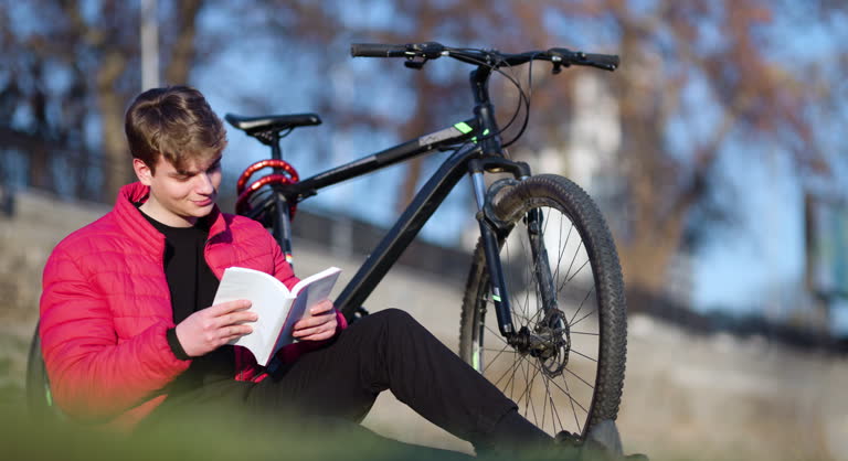 A young man relaxes with a book, seated next to his mountain bike, enjoying a sunny day in a park setting.