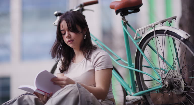 A serene image capturing a young woman engrossed in her book, seated next to her stylish turquoise bicycle against an urban backdrop.