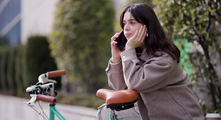 A young woman leans on her bicycle, focused intently on her smartphone, with urban foliage and buildings in the background.
