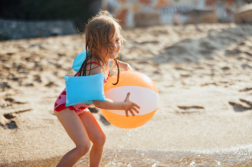 Playful young girl in a bathing suit with arm floaties, holding an inflated water ball and running into the sea on a hot summer day.