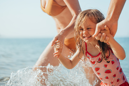 Close up shot of an adorable playful cute young girl running through sea water while holding hands of her unrecognizable parents. They are enjoying a nice sunny day at the beach. She is looking away and smiling.