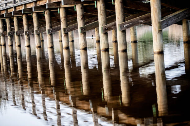 Wooden bridge over the river with reflection in the water surface. Many wooden bridges were pearls of wooden architecture throughout Europe. Unprotected wood was subject to natural degradation and decay processes. The construction elements were most often made of oak and hewn wood, over time they were replaced with resin wood, e.g. pine, fir, which better resisted the destructive effects of water.