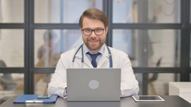 istock Doctor Smiling at Camera while Working on Laptop in Hospital 2150610114