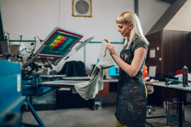 istock Print shop worker taking screen printed t-shirt off machine at facility 2150612222