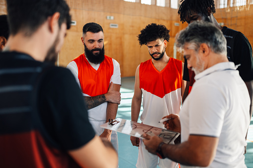 Diverse male basketball team and elderly coach in huddle while discussing game tactics. Planing the game tactic and teaching players defense. Focus on a two multiracial players. Copy space.