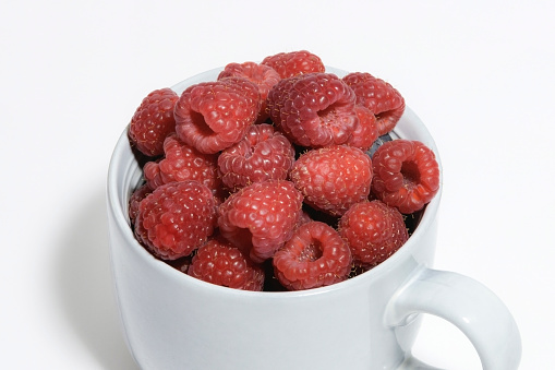 Close up of large gray mug filled to the brim with fresh raspberries on a white background