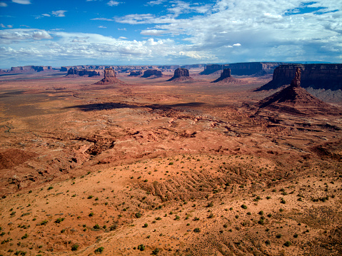 This aerial view shows a vast desert landscape with mountains in the background, located in Monument Valley near the Utah and Arizona border. The barren terrain is dotted with sparse vegetation, while the towering mountains create a dramatic backdrop.