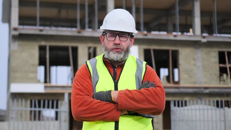 Portrait of serious engineer in hardhat crossed arms posing against building site.