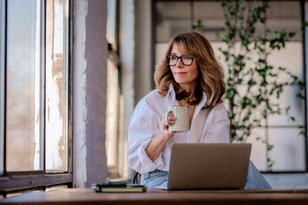 istock Attractive mid aged woman sitting at window and using her laptop for work 2150647598