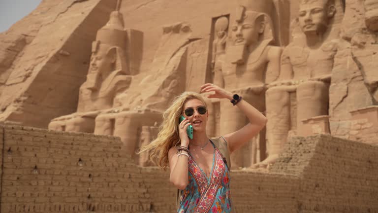 4K Video of Female in vacation talking on a smart phone standing in front of the incredible Abu Simbel Temple rebuilt on the mountain in southern Egypt in Nubia next to Lake Nasser Egypt, Aswan, Abu Simbel