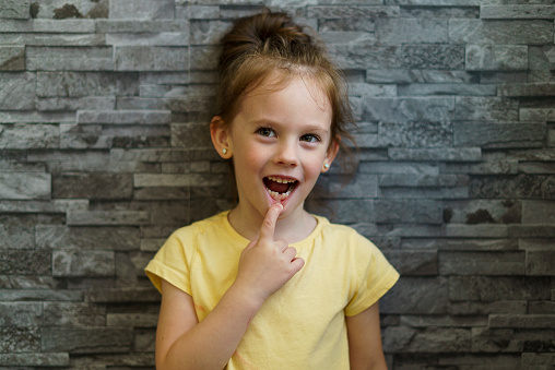 Little girl in a yellow T-shirt on a brick wall background points to a lost tooth