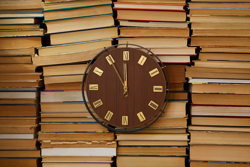 Large clock with Roman numerals on the clock face against a backdrop of stacked books. Old-school, study time, higher education, self-education